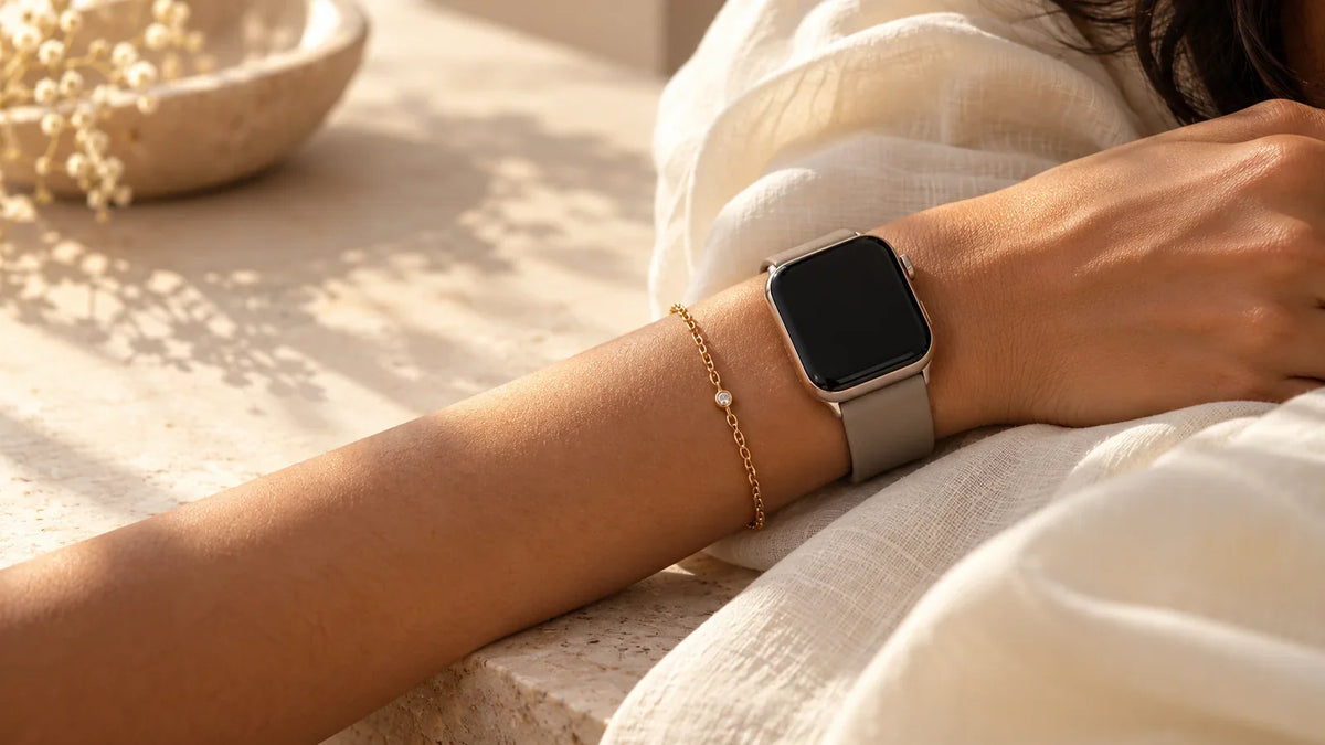 A wrist wearing a generic dark-screen smartwatch and slim gold-tone chain bracelet side by side against ivory fabric.