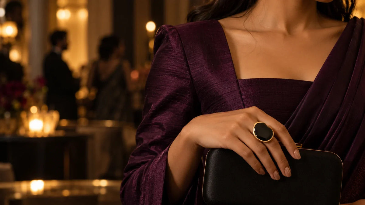 Conceptual editorial: a sculptural black-and-gold-tone ring on a hand in a plum evening sleeve, above a black clutch; not an ARVENIER product photograph.