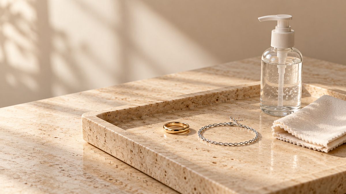 A gold-tone ring and silver-tone bracelet beside an unlabelled sanitiser bottle and soft cloth on a stone tray; conceptual still life, not an ARVENIER product.
