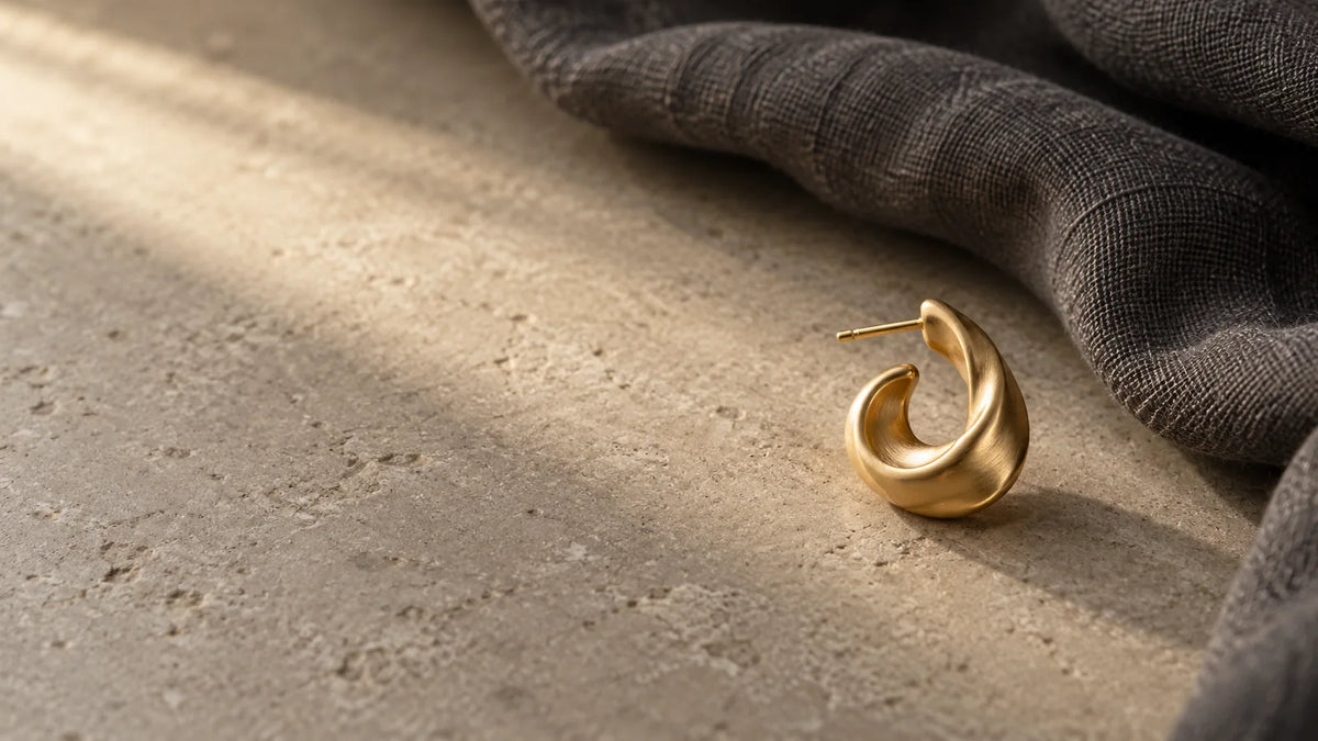 Conceptual editorial still life of a single sculptural gold-tone earring on matte stone beside charcoal linen; AI-generated, not an ARVENIER product photo.